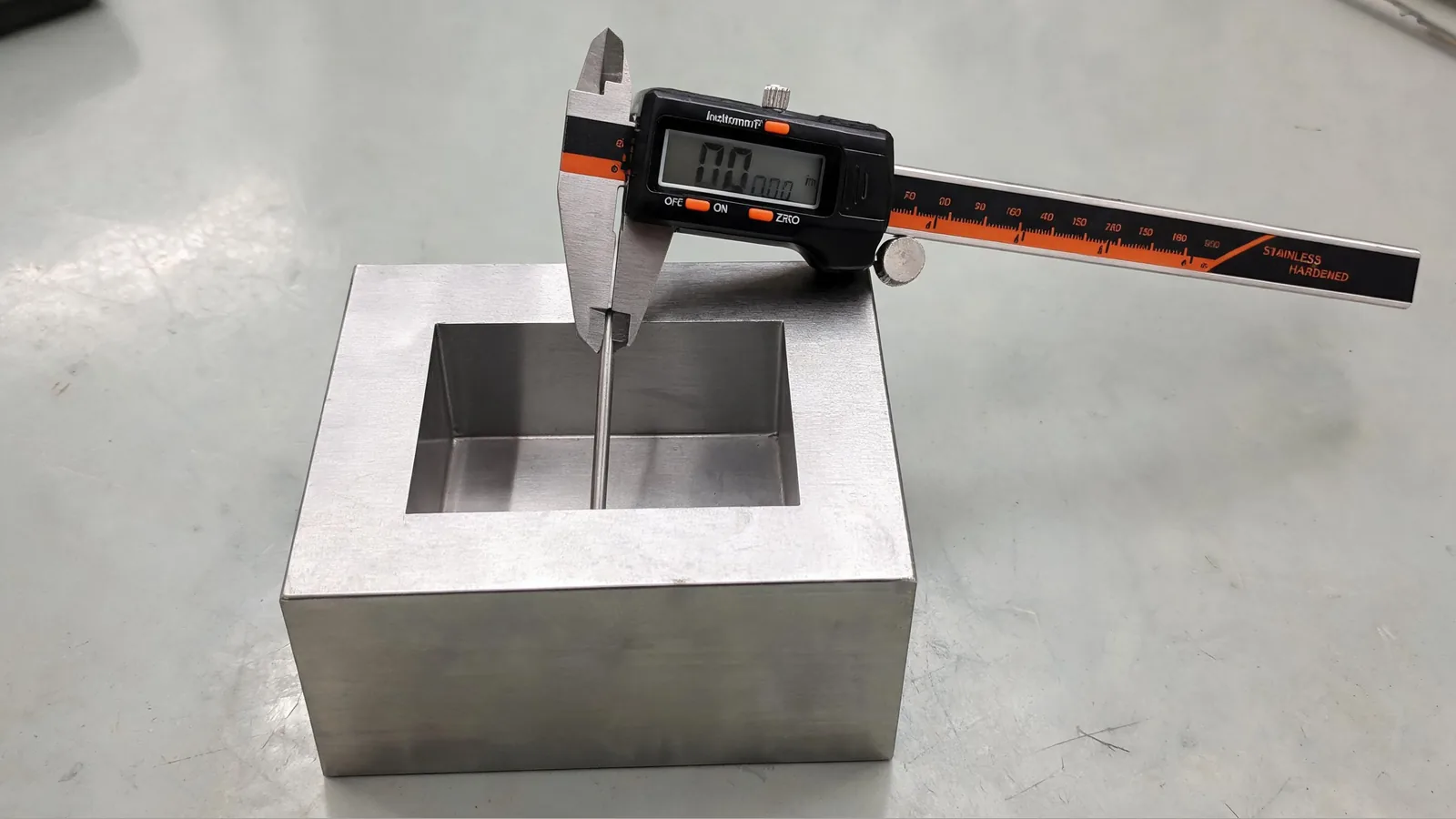 Digital caliper measuring the depth of a square metal recess, with the beam resting flat on the top surface and the depth rod extended vertically into the cutaway.